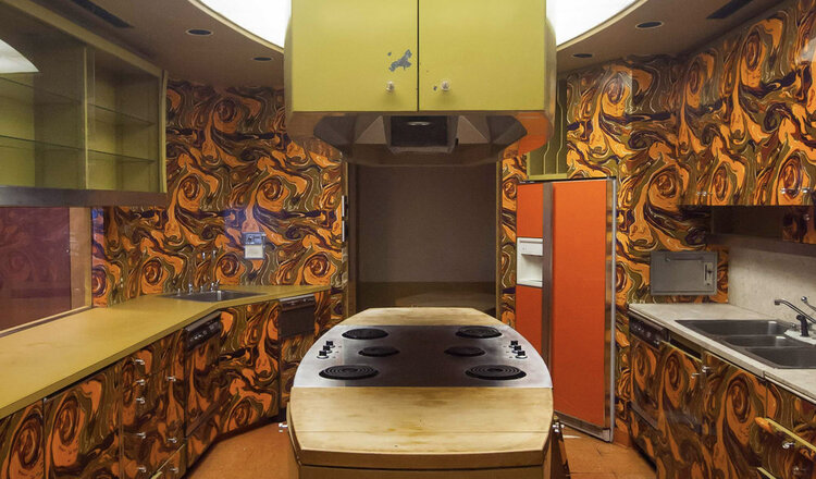a kitchen with swirly orange and brown wall paper, lime yellow cupboards, an orange fridge, and wooden countertops