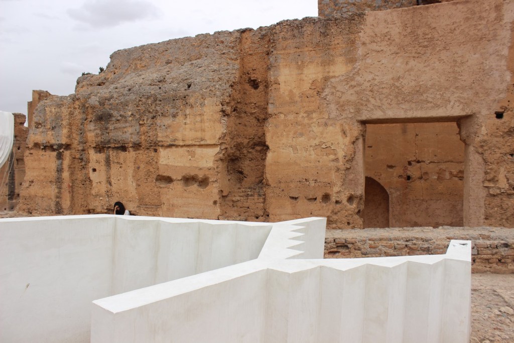 a  white cement sculpture of stairs on its side, photographed in front of ruins of red stoned buildings - the photograph shows the stark contrast between the two objects