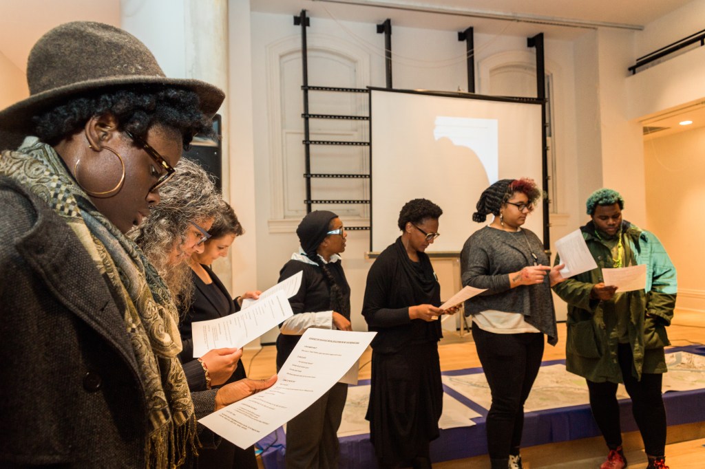 a variety of people stand in a semicircle with papers in hand, reading aloud