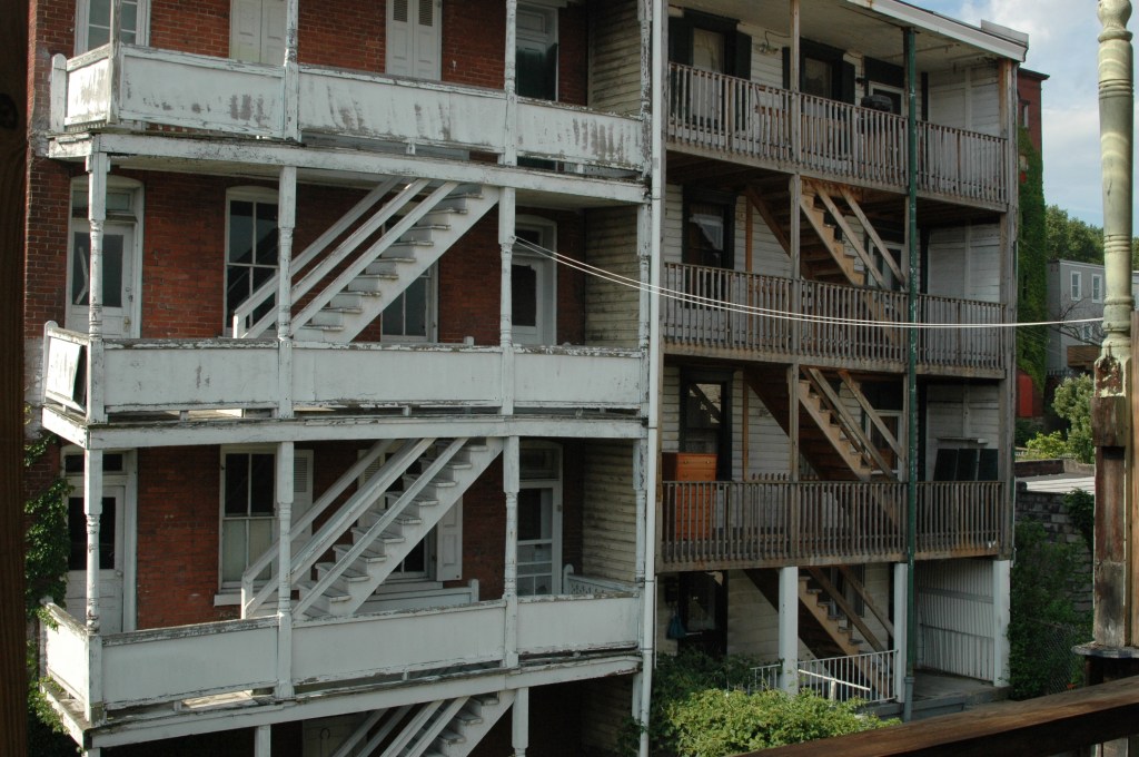 a view from the Backyard of Srscott's house - there are two four-story buildings with porches and connecting stairways. Both buildings are brick with wooden stairs and railings, and they paint on them is peeling