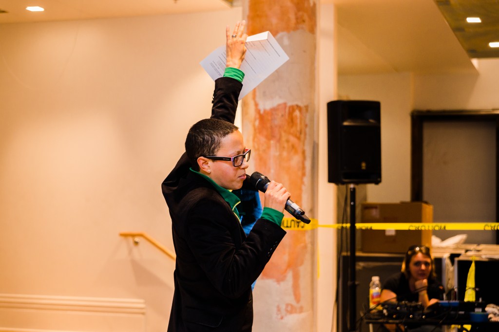 Mitch, a Black woman with shaved hair, thick Black framed glasses, and a suit stands with a pic in one hand with the other hand raised high up in the air