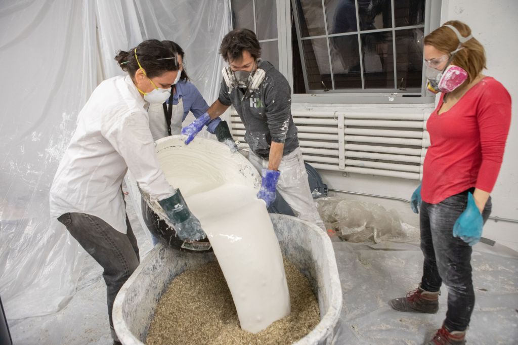 Jonsara Ruth and three of her colleagues stand on a plastic drop cloth wearing gloves and face masks. They are dumping a large bucket of lime mixture into a vat of dried hemp leaves to make Hemplime.