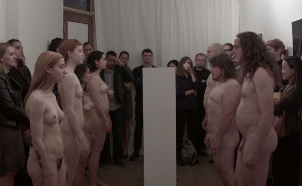 7 nude performance artists lining either side of a blank white wall, staring at it, surrounded by a crowd of people