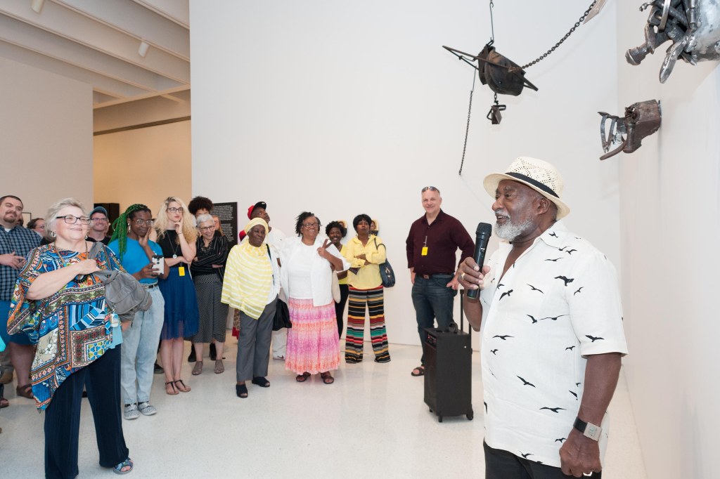 Mosley stands in w white short sleeve button down with a bird pattern with a white fedora in a gallery in front of a crowd, explaining his art piece