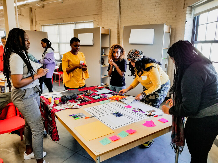 a group of black women gathering around a table completing a workshop exercise with markers, sticky notes, and posterboard