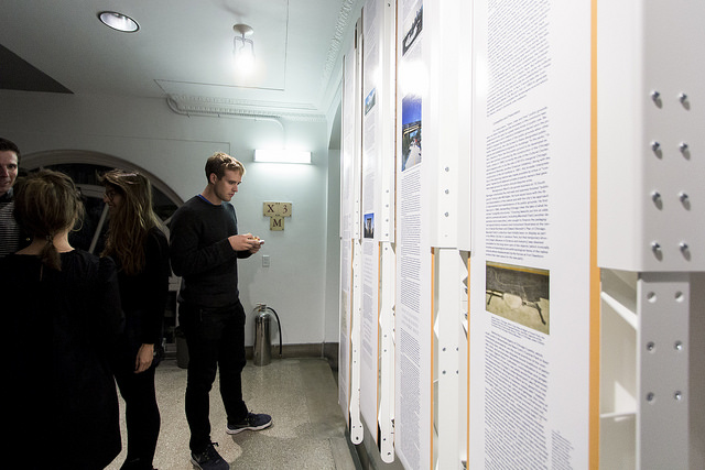 a white industrial looking, metal gallery wall full of text with people standing in front