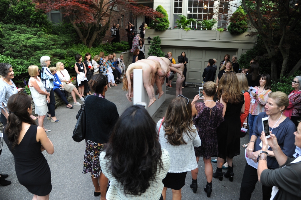 5 nude performance artists, draped over a white 5 foot tall blank wall outdoors, surrounded by a group of people