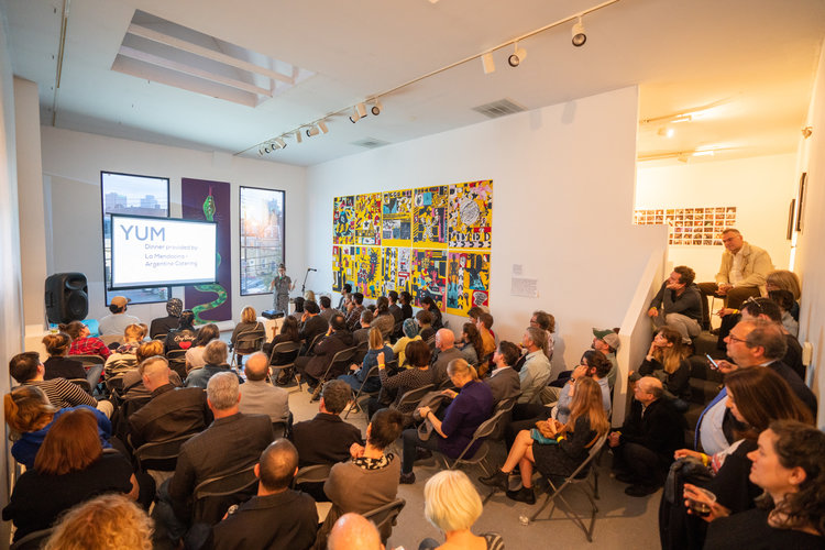 a packed presentation space with people sitting in folding chairs and on stairs adjacent to the space - on one wall is a presentation screen, on another is a variety of photos