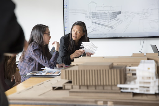 anne chen holding a white model, discussing with someone seated next to her. In the foreground is a brown chipboard model of Hillman Library, a large, square concrete building