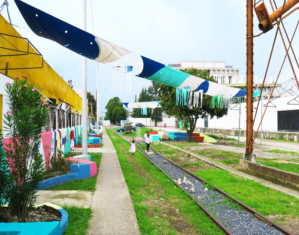 an old railroad turned into a park -bright green grass grows on either side of the track, fabric is suspended above providing shade, and a bright wall next to an adjacent sidewalk is painted with colorful murals
