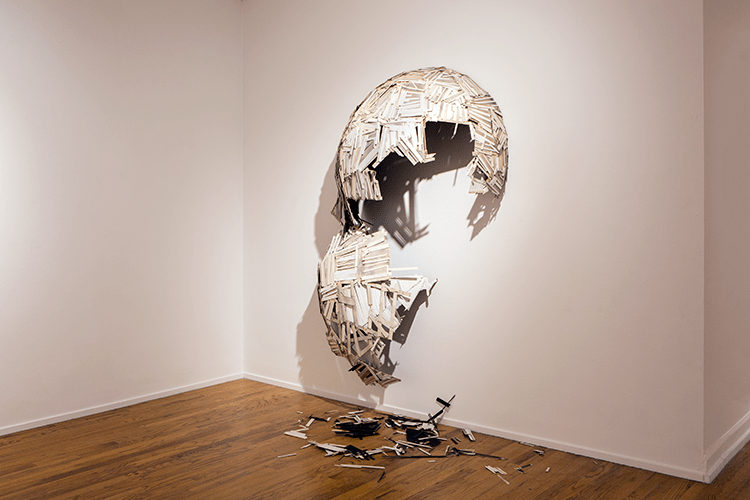 a dome on a gallery wall made of wooden sticks - the dome is white and the wood of the sticks is still visible. The dome is broken, half of it hangs from the wall, and some of the material from it lies on the floor below. 