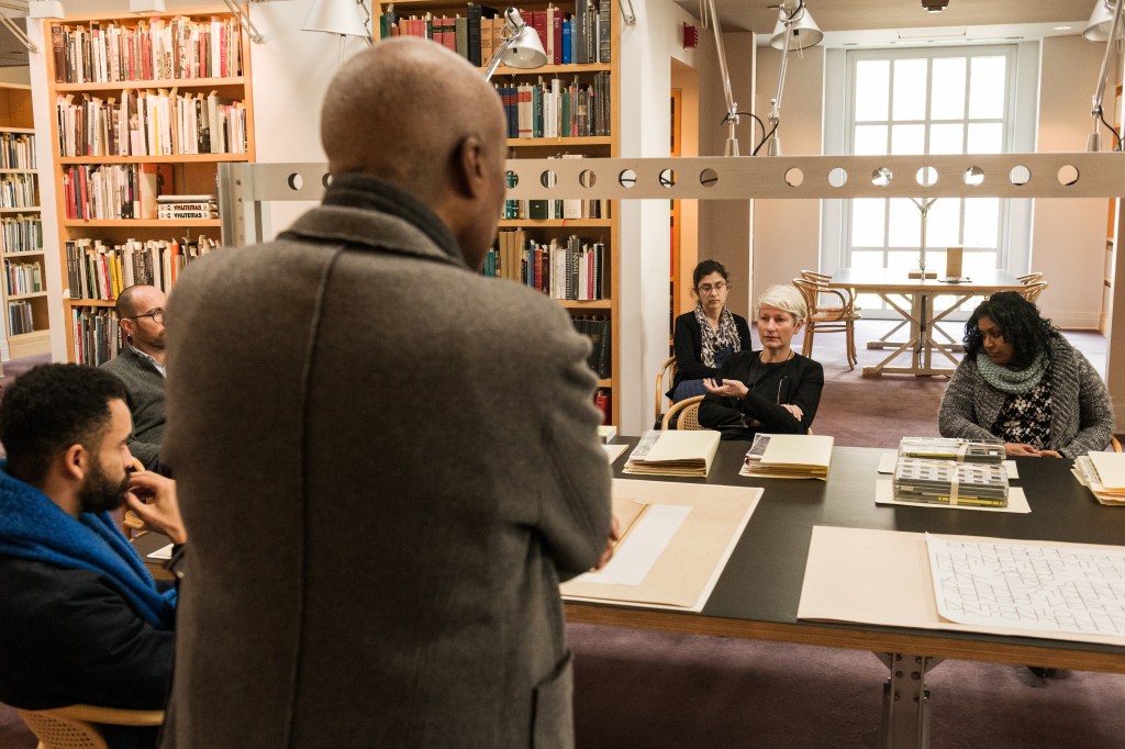 Giovanna sits with arms crossed at a table full of large archival documents in a library. People sit in chairs around here, and a man is standing in the foreground attentively listening to her