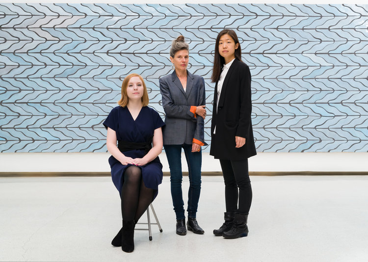 Liz Park with short red hair, sitting in a blue dress, Ingrid standing center in a gray blazer, and Ashley McNelis standing in a black cardigan - all in front of a pale blue tiled art piece