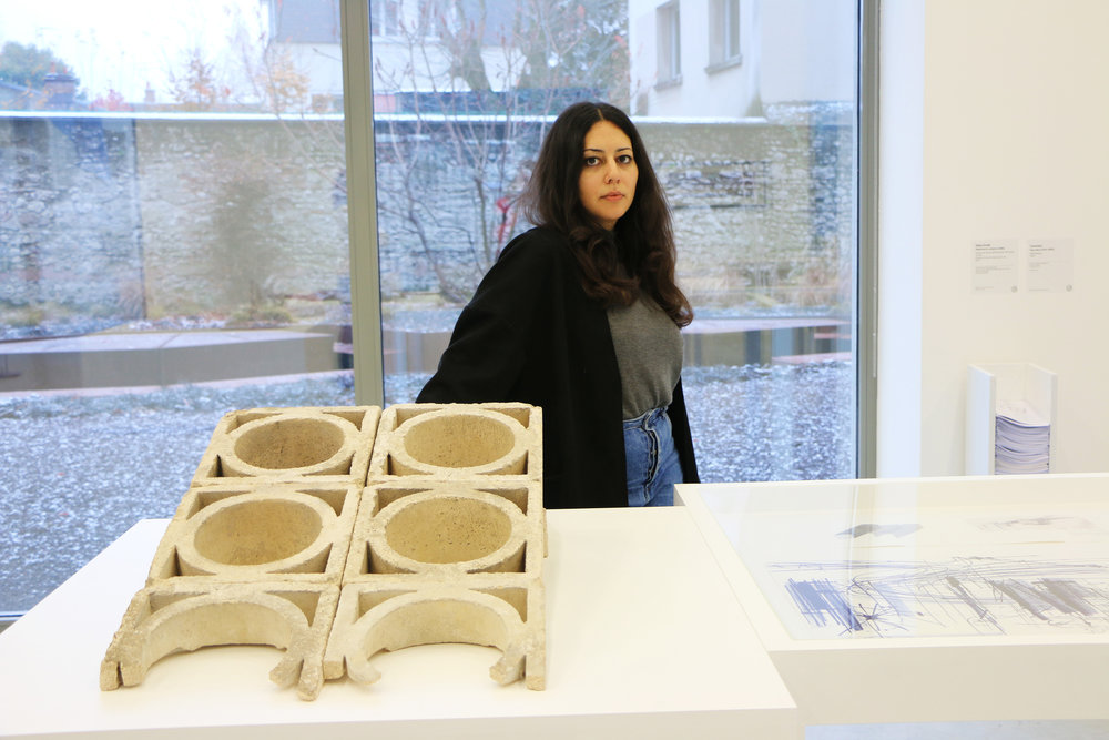 a photo of Saba Innab - with long dark hair, wearing a black cardigan and a gray shirt. She stands in front of a building fragment.