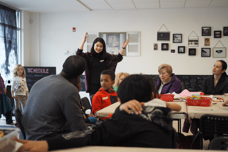 Annie, wearing a sweater and with long dark hair, with arms in the air, leading a workshop in a room with people of all ages sitting around tables full of art supplies in small red baskets