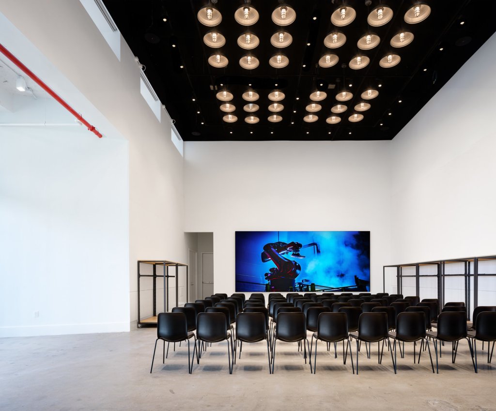 a white room with a high black ceiling with circular lights - in front of a large projector screen are rows of black chairs