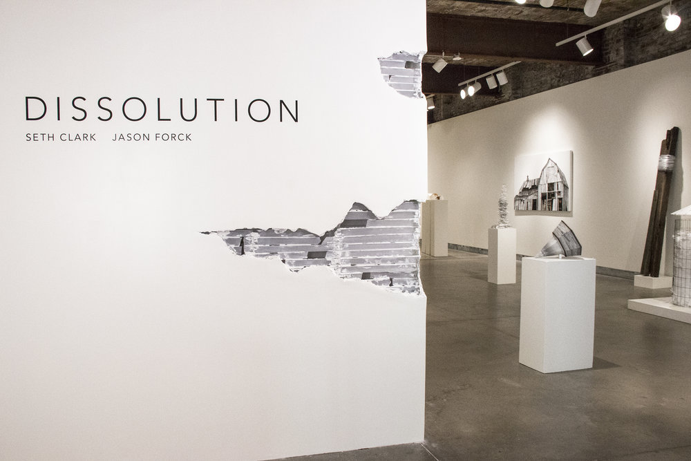 a white gallery wall with the title of the exhibition: Dissolution, in black text. the wall is slightly decayed, with two spots with plaster falling off.