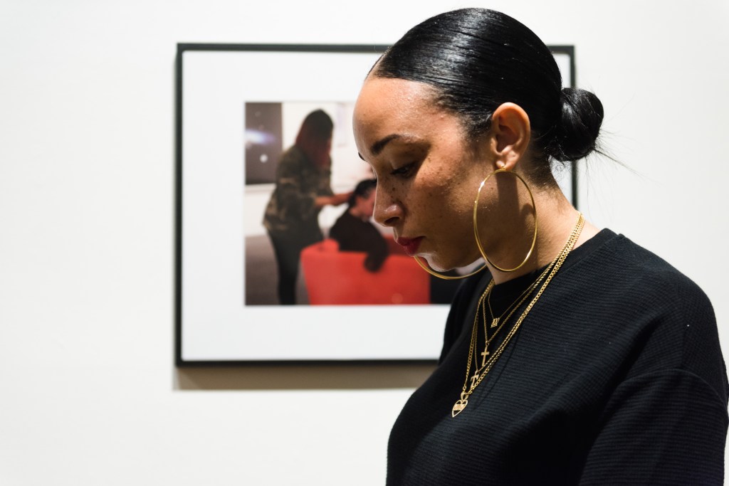 Tara with her hair tied back in a low bun, wearing gold necklaces and hoop earings, in front of a photo of her performance piece where she is getting her hair braided