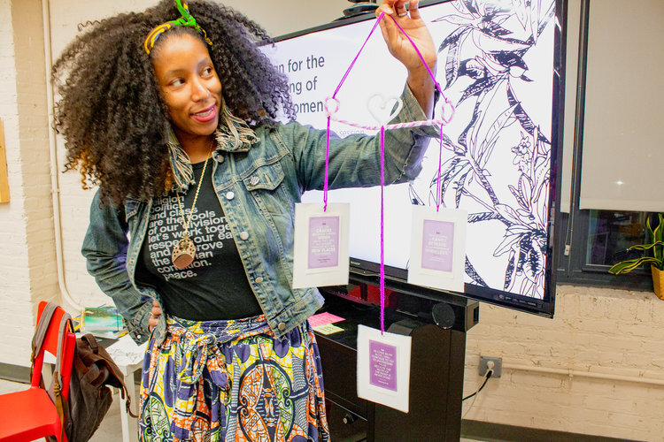 a black woman with a beautiful fro holds up a pink mobile she made