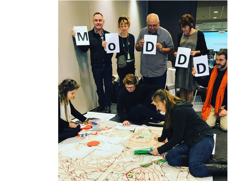 a group of people standing and holding up pieces of white paper with black letters that spell out MoDD. Below them are people on the ground, looking at a large scale map.