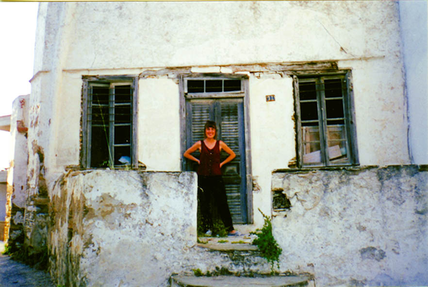 Theocharopoulou, with hands on her waist, smiling in front of new new home - plaster is chipped and falling, the paint on the door and window frames is peeling, and plants are growing in the cracks of building - it looks like the space hasn't been touched in years