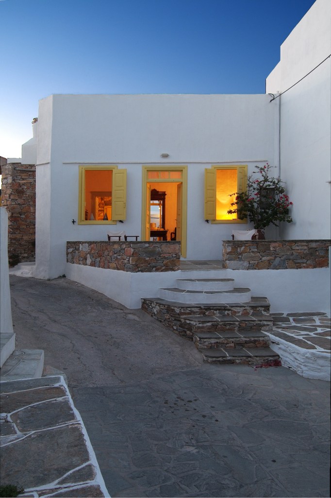 a drastic change from the previous image where it looked like the building wasn't touched in years, the exterior is now smooth and repainted in white, the door and window frames are painted a bright yellow, and a stone railing has been added. warm light pours from the interior, and it looks nice and home-y