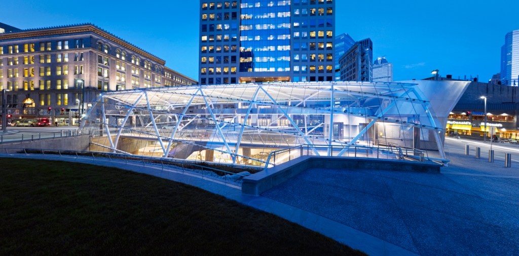 a steel and glass structure that arches over and covers the underground entrance to the train - the glass reflects the blue hue of the sky