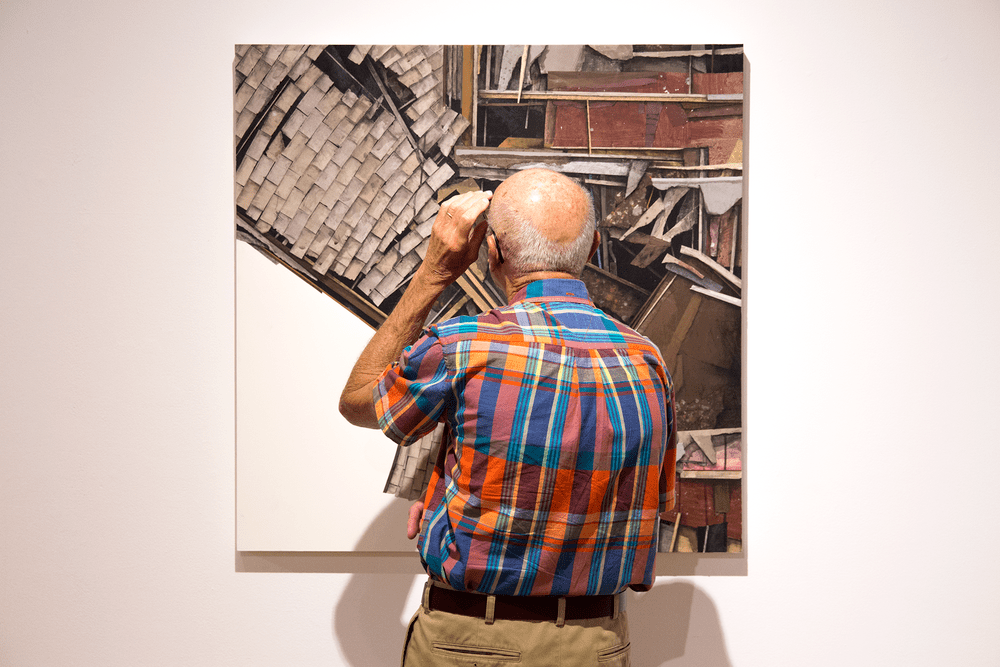 a gallery visitor in a colorful plaid shirt looks up close at one of Clark's artworks - a collage of different materials like roof shingles and window frames
