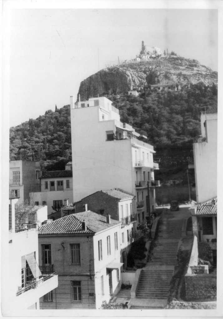 a black and white image, aged at the edges, of a white rectangular building shaped like steps near the upper floors. the building rests on an incline with 3 sets of stairs in front of it