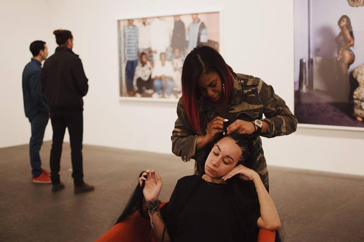 Tara sitting in a red armchair in the middle of a gallery, getting her hair braided by Shaquille Douglas, who has black and red ombre hear and is wearing a camo print shirt. Behind them are two photographs capturing black folks.