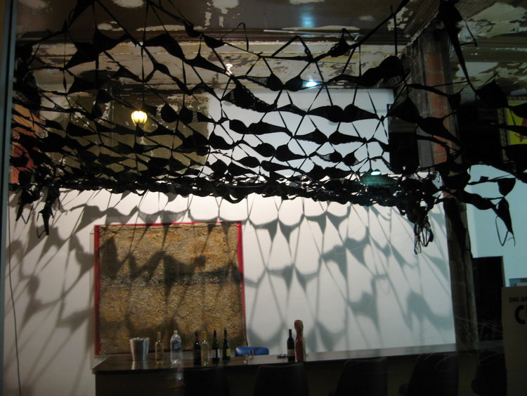 an installation that looks like a black woven net - a light from above casts a unique pattern on an adjacent white wall