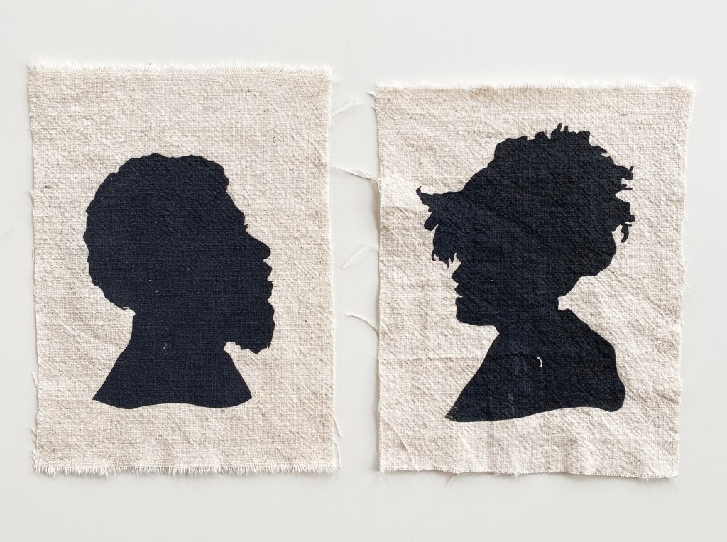 two black silhouettes - one of a man and on of a woman - are on two pieces of white textile. The two silhouettes face each other, almost like they're talking to each other