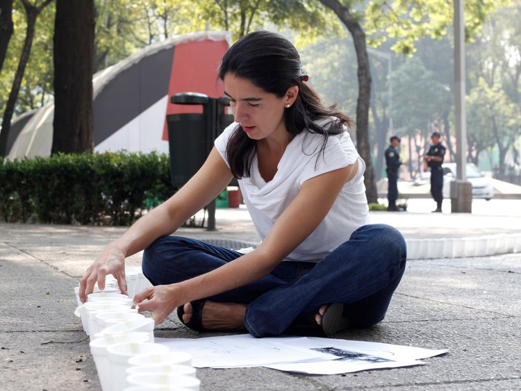 Fernanda Canales in a white t shirt and jeans, hair down, sitting on the ground outdoors with papers and working with a row of white Styrofoam cups