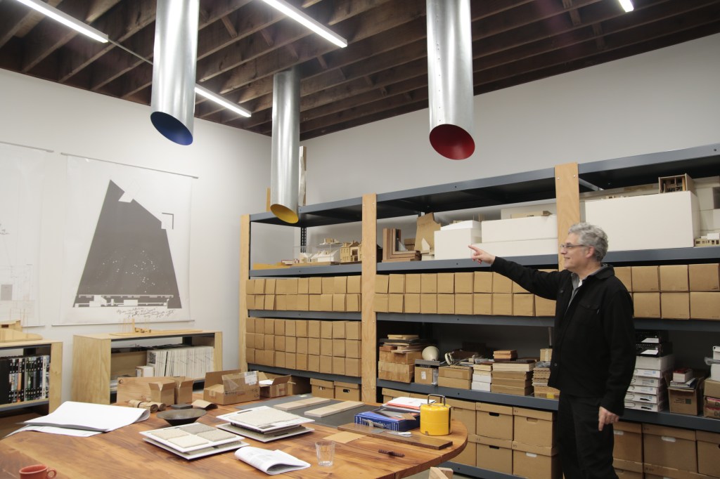 Gerard standing and pointing at some drawings in his studio - a space with white walls and shelves stocked with small organized cardboard boxes and a table with material samples. Three primary colored lamps come down from the ceiling.