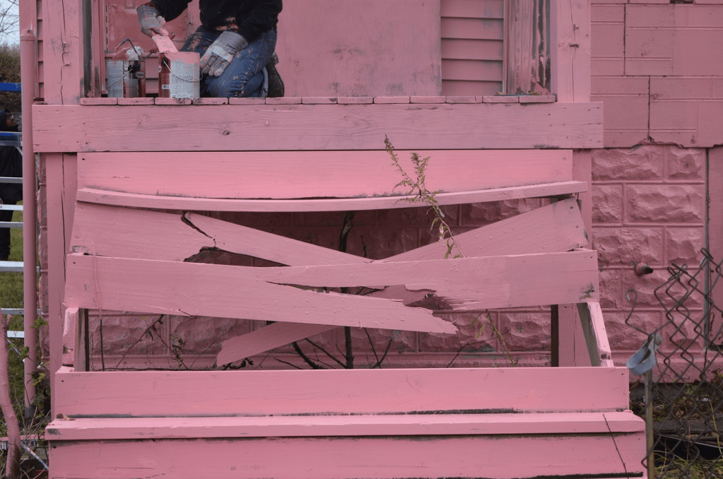 steps leading up to a house, splintered and broken, being painted pink (the color of Pink Oil Moisturizer) by someone sitting at the top of the stairs with multiple buckets of paint and a brush