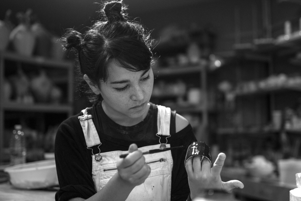 a moody, black and white image of Diedre Shea - Shea has their hair in two buns and is wearing overalls and working on painting a ceramic piece