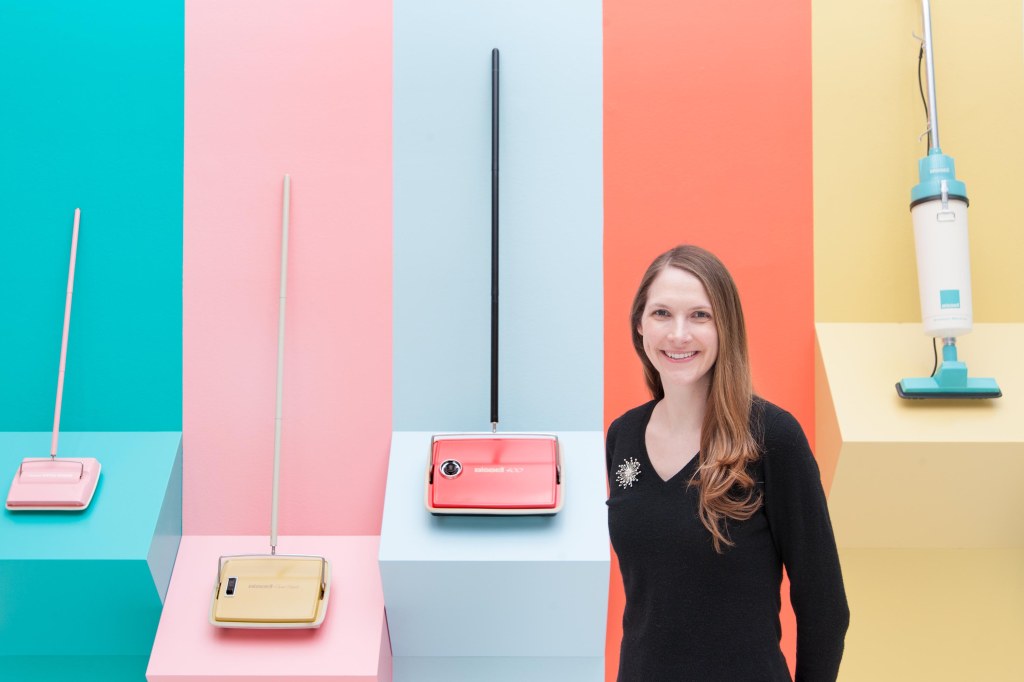 Rachel, with long brown hair in a black sweater, standing in front of a pastel wall sectioned off to display pastel push vacuums 