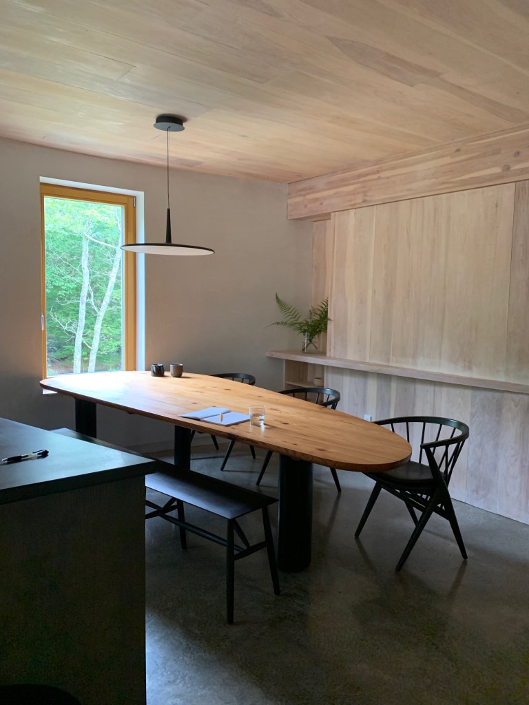 An oblong wooden dining table nestled in a corner by a window. The table is flanked by three black wooden dining chairs with deep-set rounded backs. Behind the table is a floor to ceiling wooden shelf system that is light beige in colour. The walls are finished in white lime plaster. The floor is a dark grey lustrous concrete.