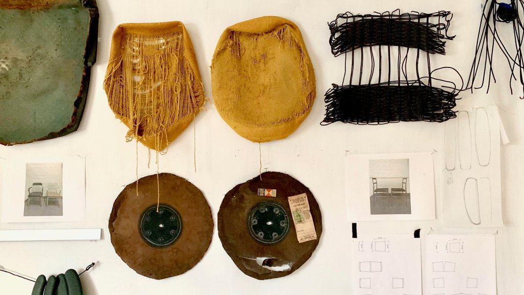 A white bulletin board covered with pencil sketches, printed photos of chairs, and pieces of old mid-century modern furniture, including  a green and brown cushion cover, two fraying ochre chair seats, and a black woven seat with holes in it.