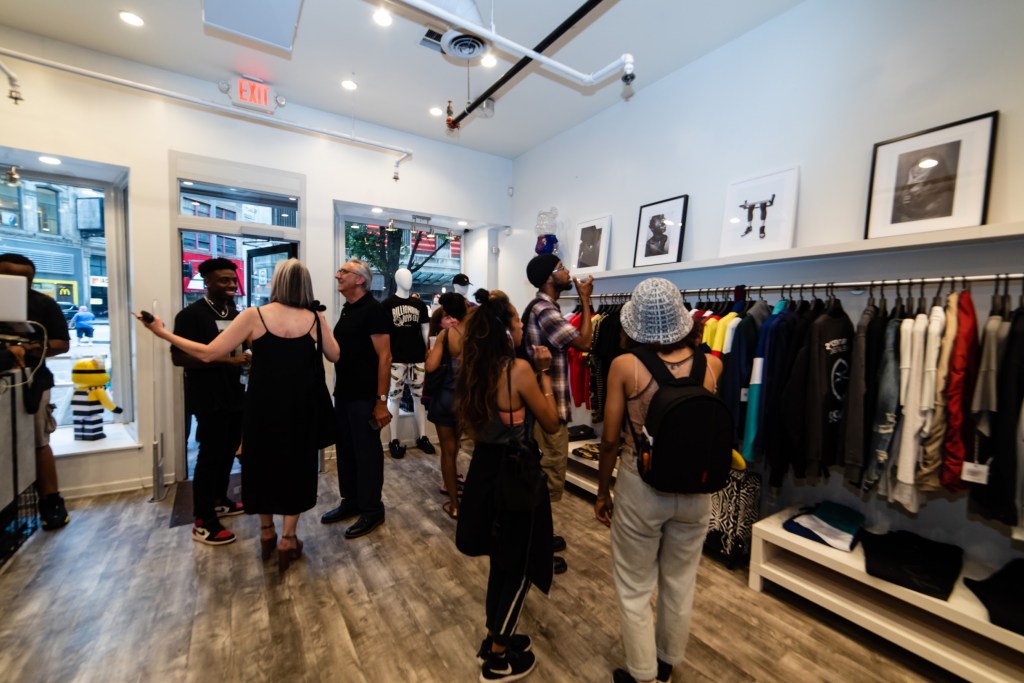 a group of people standing in a storefront, with black and white art pieces propped up on a shelf above a rack of clothing