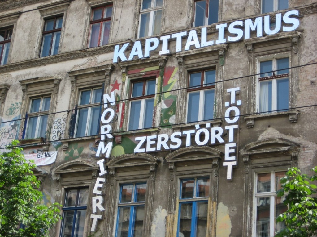 an old brick facade with a message in German in white block letters that reads: Capitalism normalizes, destroys, kills 