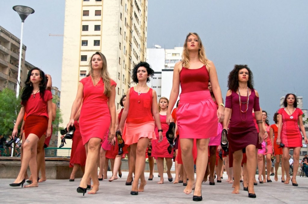 a group of women in various shades of pink and red, all wearing one black high heeled shoe
