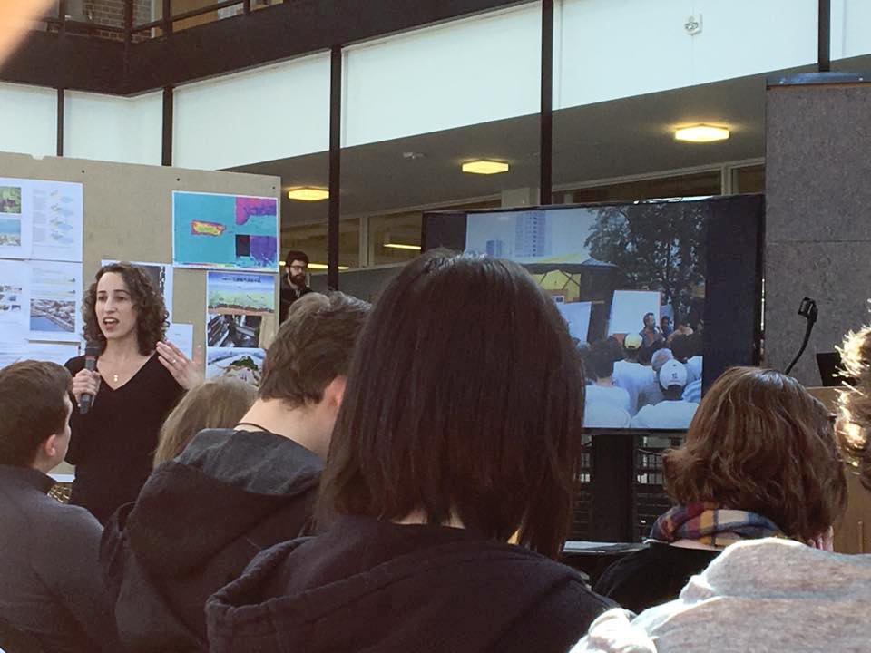 sandler, in a black shirt with brown shoulder length curly hair, stands in front of a full room with a tv monitor behind her, mid-discussion