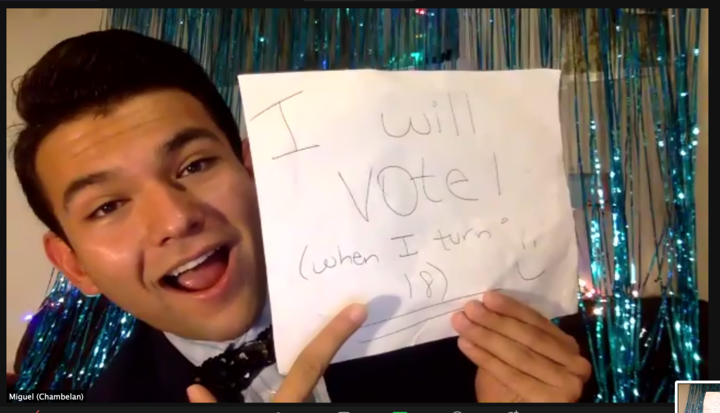 a young man wearing a tux holds up a handwritten sign that says "I will vote! (when I turn 18)" - he's smiling and pointing at the sign enthusiastically 