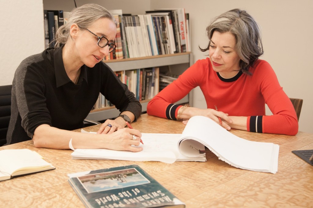verona, in a black collared shirt, and carpenter, in a red sweater, sit at a table  and are captured mid discussion, with verona sketching something on trace paper