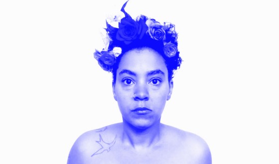 Alisha pictured against a stark white background, wearing a flower crown of pink and red roses in their curly hair with a bird tattoo on their left shoulder. They're staring straight into the camera.