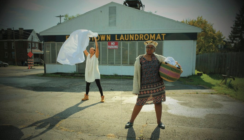 Two womxn stand in front of a laundromat in an empty lot. In the foreground, one stands with a powerful stance with a woven basket full of laundry resting on her hip staring into the camera. In the background, another womxn is flapping a white sheet, airing out laundry. The image has a black faded edge, making it look like an old, archival image.